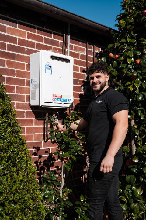 An All Day plumber beside a Rinnai hot-water heater at a customer's home.